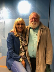 The image shows Miriam O'Callaghan and Frank McGuinness side-by-side. Miriam is on the left, perched on a seat. She is wearing a blue blazer and has long blonde hair. Frank is standing to the right. He appears to have his arm around Miriam. He is wearing a pastel green shirt with a light brown overcoat. He is bald with a bushy grey beard.