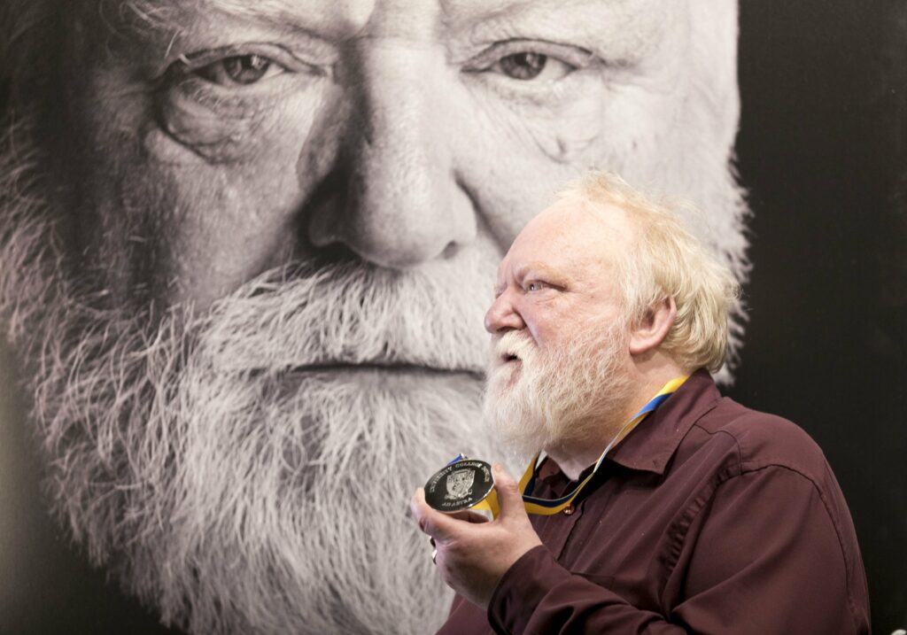 The image shows Frank McGuinness in profile holding up a medal. He is standing in front of a large-scale black and white photograph of himself.