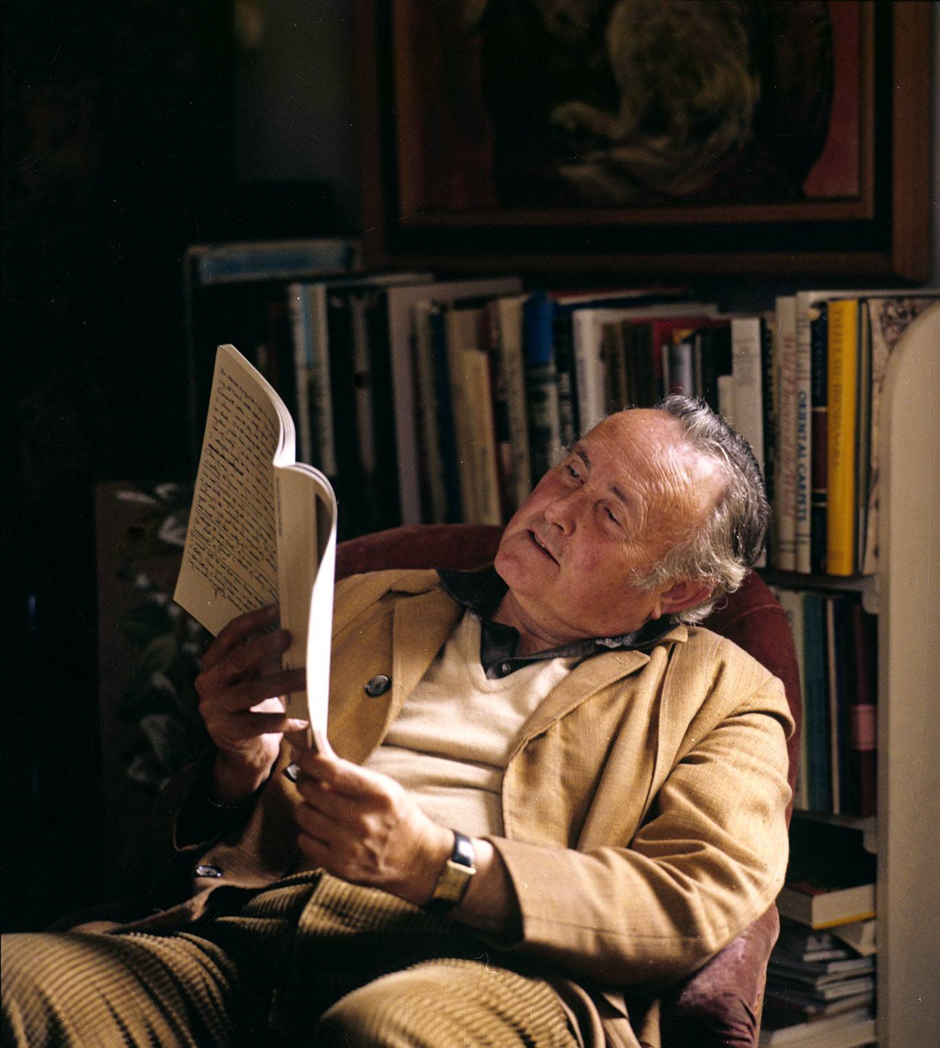 The photograph shows artist Derek Hill reading a book. He is sitting in an armchair in front of a bookcase. His head is tilted sideways as he directs the pages of the book to catch the light.